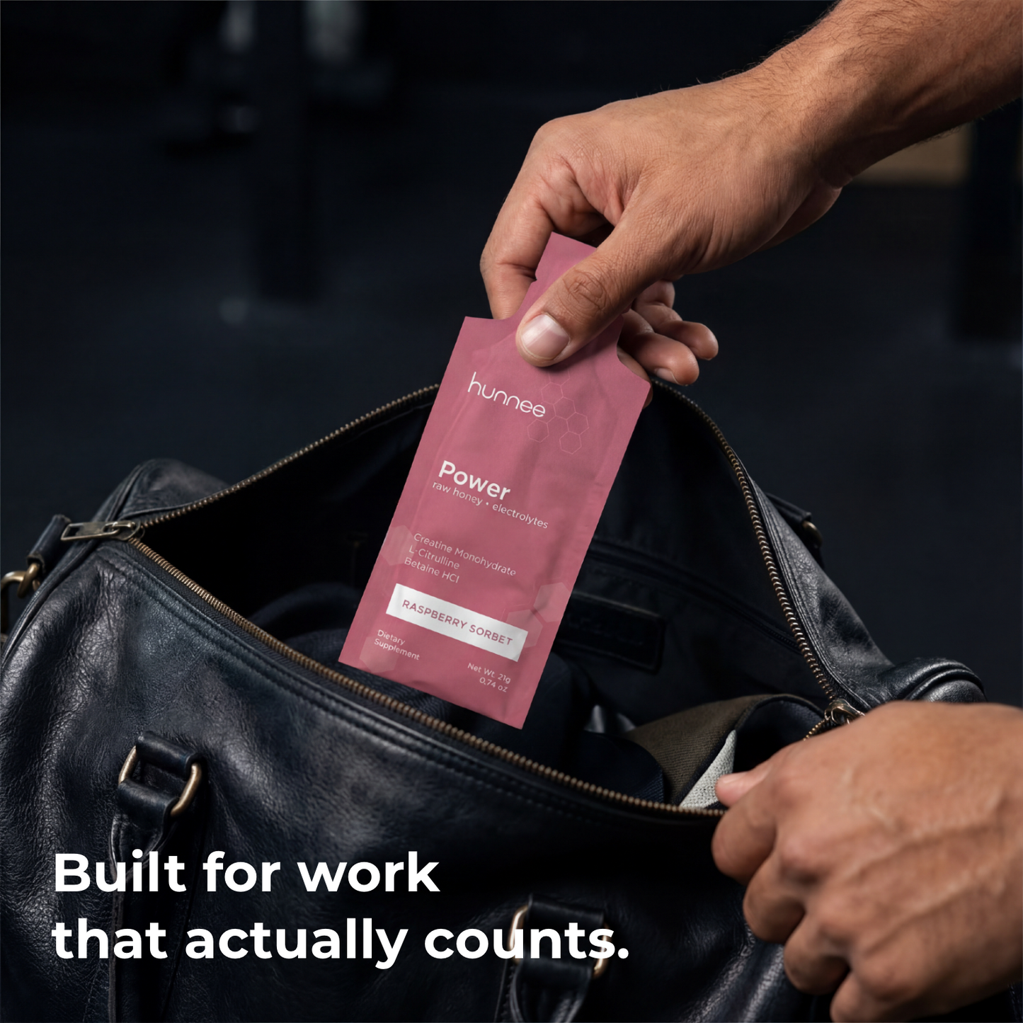 Person holding a pink 'hunnee' product package in front of a black leather bag with text 'Built for work that actually counts.'.
