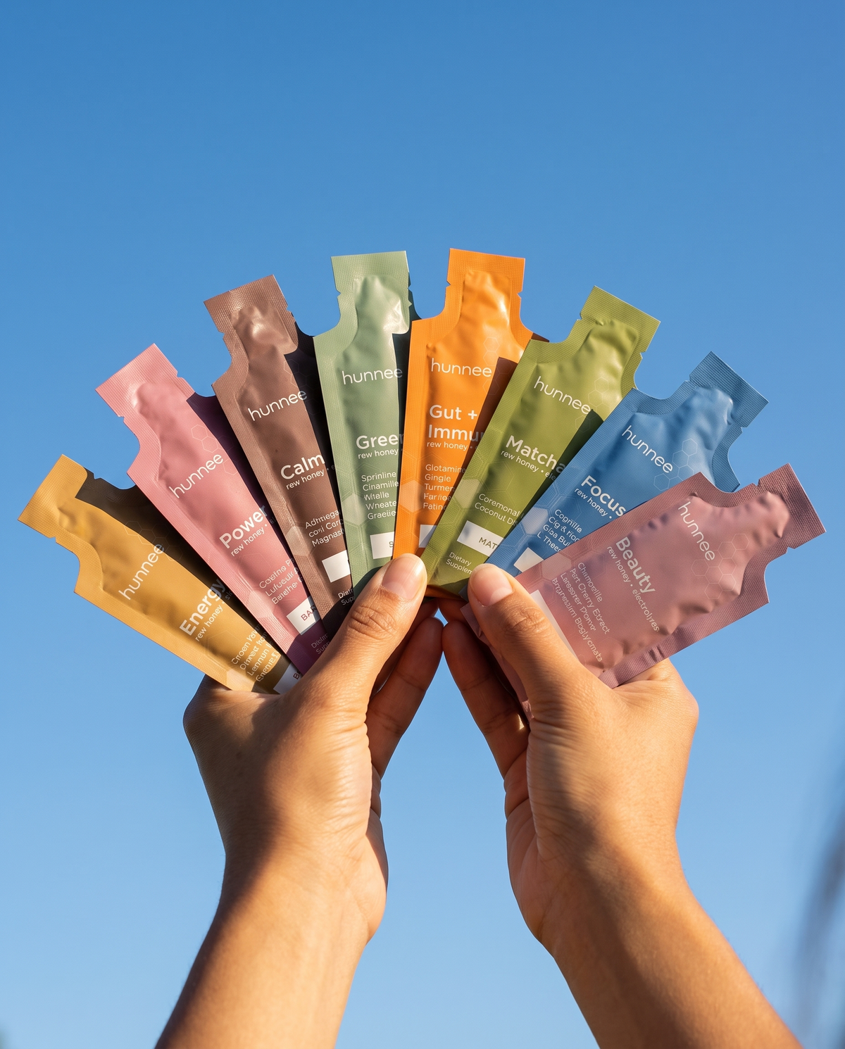 Hands holding a fan of nine hunnee superfood honey sachets against a blue sky